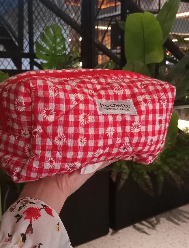 Red gingham cotton beauty case with white floral print, held by hand, indoor plants background