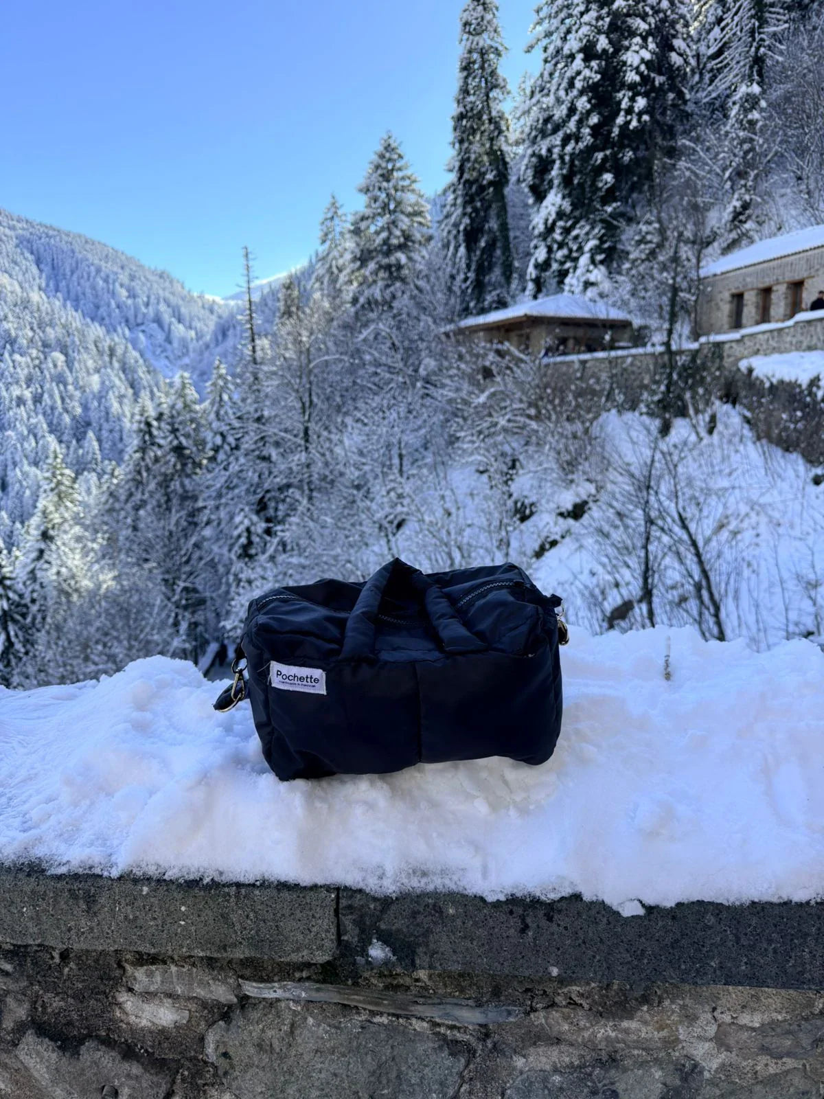 Black puffer quilted crossbody bag on snowy ledge with winter forest and cabins in background