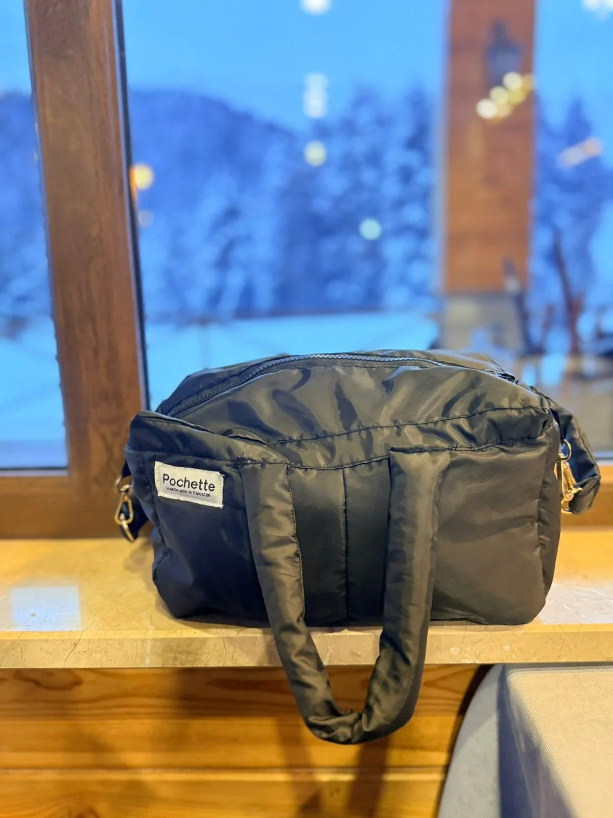 Black quilted puffer crossbody bag with handles on a windowsill, snowy background outside.
