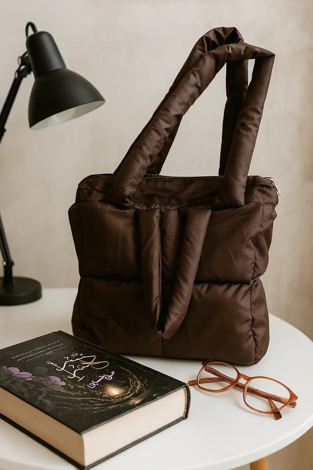 Brown cotton padded tote bag on a white table with a book, glasses, and desk lamp